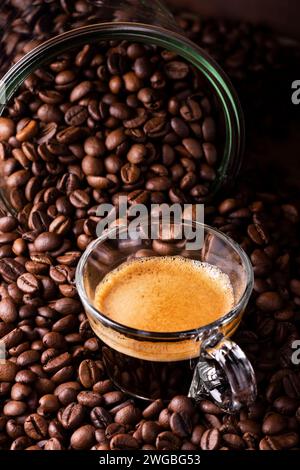 in the foreground a glass cup with espresso coffee. The cup is surrounded by roasted coffee beans. Stock Photo