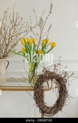 Close-up of a bunch of yellow tulips in a vase on a table with a rustic wreath and jug with branches Stock Photo