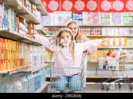 Cheerful cute girl sitting in a shopping cart at the supermarket, her mother is pushing her: grocery shopping and lifestyle concept Stock Photo