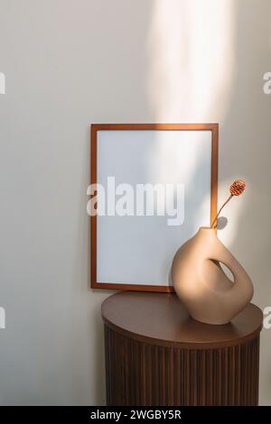 Blank wooden picture frame and a ceramic vase on side table against a wall with dappled light Stock Photo