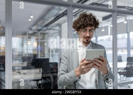 A serious and focused young man is standing in an office center and working on a project using a tablet. Stock Photo