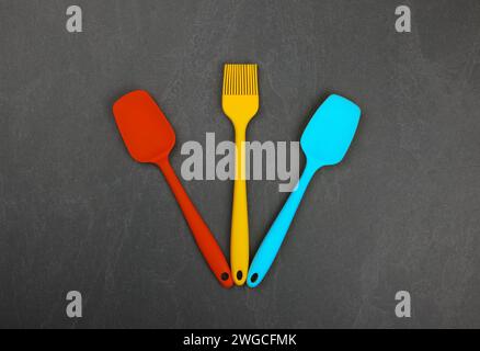 Three silicone cooking spatulas and brushes on black slate board with copy space, colorful modern culinary kitchen utensils, close up, elevated table Stock Photo