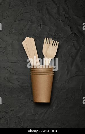 Close up disposable brown paper takeaway coffee cup, wooden spoons and forks on black background, natural eating and drinking utensils, close up, elev Stock Photo
