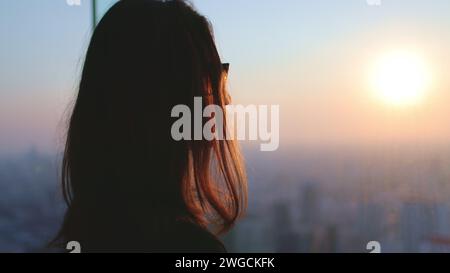 Woman enjoy cityscape sunset aerial view standing near the window. Close up long hair girl silhouette in sunglasses against bright colorful sky, sun goes down, urban skyline background Stock Photo