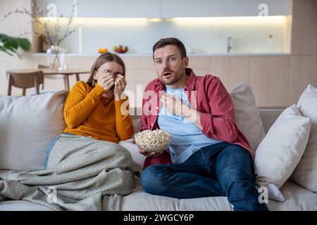 Scared afraid fearful family couple watching thriller horror eating popcorn sitting on home sofa  Stock Photo
