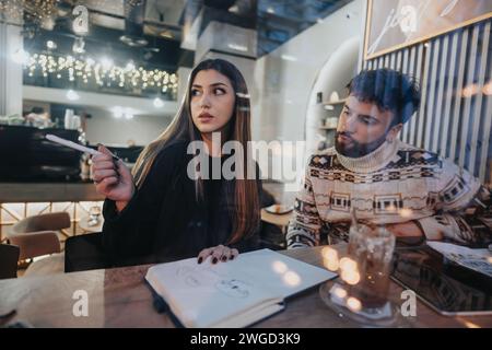 Two creative young people are engaged in a collaborative brainstorming session in a warmly lit cafe, with focus on teamwork and productivity. Stock Photo