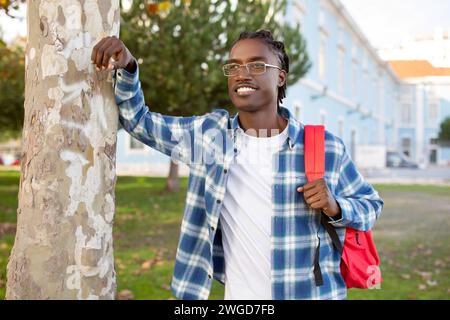 Cheerful black student guy with backpack leaning against tree outside Stock Photo