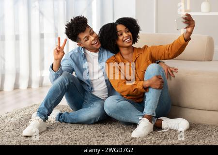 Sitting comfortably on the floor, a delighted young couple enjoys a light-hearted moment taking a selfie Stock Photo