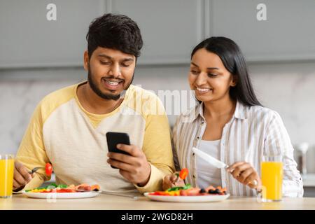 Millennial Indian Man And Woman Having Breakfast And Using Smartphone Stock Photo