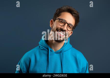 Smiling young man wearing glasses and bright hoodie standing against blue background Stock Photo