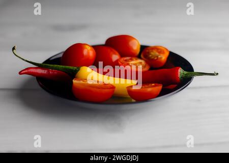 One and a half hot peppers on a black round plate. Cherry tomatoes. Light background. White boards. Spices in the kitchen. Stock Photo
