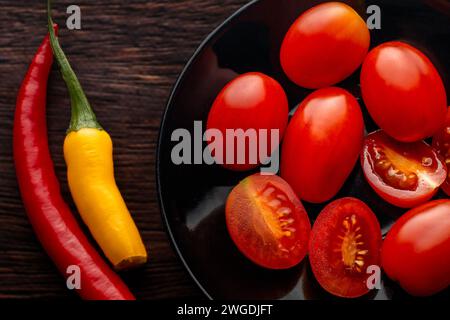 Cherry tomatoes, red and yellow peppers on a black round plate. Dark background. Brown boards. Spices in the kitchen. Stock Photo