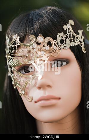 Plastic woman mannequin with black hair wearing a golden half mask with diamond gems on a blurry trees background outside in natural light Stock Photo