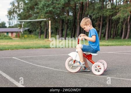 Very focused Little boy in blue, riding a red old tricycle in city park. Stock Photo