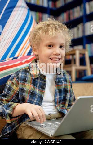Vertical portrait of cute blonde boy with curly hair using laptop in school library and smiling at camera Stock Photo