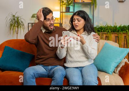 Disappointed multiethnic couple reading together bad news on smartphone while sitting on sofa in room at home. Sad Hispanic family are depressed by unpleasant message shocked by information about loss Stock Photo