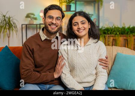 Loving young diverse couple looking at camera and smiling sitting arm around relaxing on sofa. Happy Indian family spending leisure time together on couch looking at camera in living room at home. Stock Photo