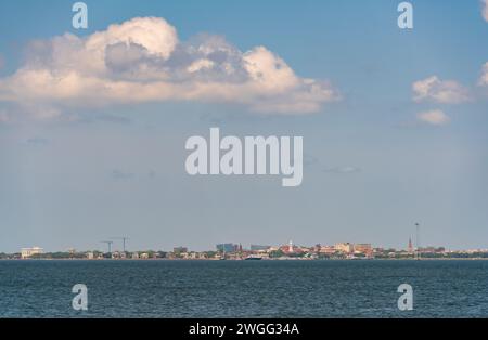 A large white cumulus cloud floats over a thin line of distant buildings and cranes above deep blue harbor water in Charleston, South Carolina. Stock Photo