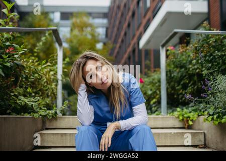 Young female doctor feeling overwhelmed at work, sitting on stairs, looking at phone. Healthcare workers having stressful job, feeling exhausted Stock Photo