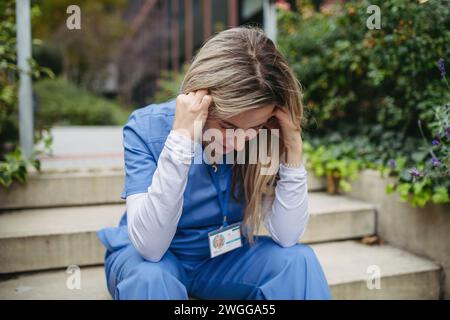 Female doctor feeling overwhelmed at work, sitting on stairs. Healthcare workers having stressful job, feeling exhausted. Burnout syndrome for doctors Stock Photo