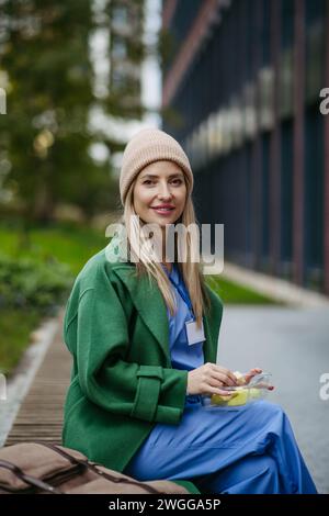 Nurse having healthy snack in front of hospital building, taking break from work. Importance of breaks in healthcare. Stock Photo