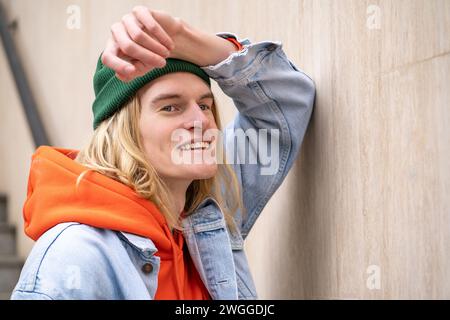 Carefree long-haired hipster guy wearing trendy stylish outfit, stand in city street leaning on wall Stock Photo