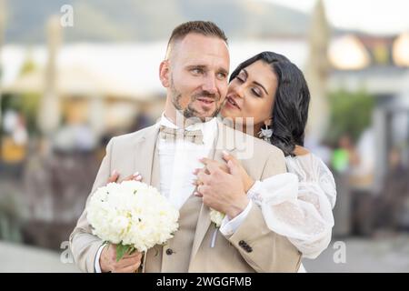Wedding portrait of smiling newlyweds. A stylish groom in a beige suit and a cute brunette bride in a white dress are tenderly embracing in the park. Stock Photo