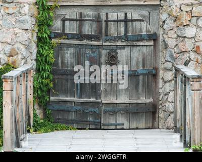 Old wooden gate with forged hinges in a stone wall overgrown with ivy and a bridge with wooden railings in front of it Stock Photo