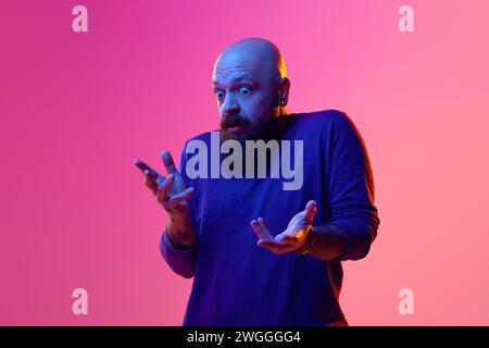Portrait of bearded bald man in swear expressing emotions, standing with shocked face against pink background in neon light Stock Photo