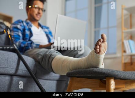 Young man with broken leg sitting on sofa using laptop Stock Photo