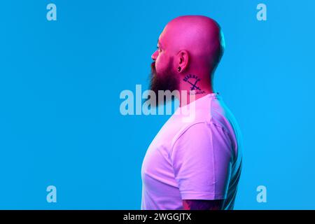 Portrait of bearded bald man with tattoo on neck, in casual white t-shirt posing against blue background in neon light. Side view Stock Photo