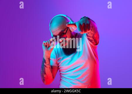 DJ. Bearded, artistic bald man with tattoos, in white t-shirt and sunglasses standing against gradient purple background in neon light Stock Photo