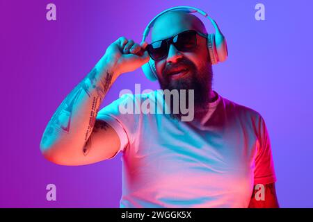 DJ. Bearded, artistic bald man with tattoos, in white t-shirt and sunglasses standing against gradient purple background in neon light Stock Photo