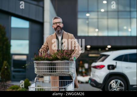 Man charging electric car before going to shopping. Public charging station in front of shop building, supermarket. Charging while shopping. Stock Photo