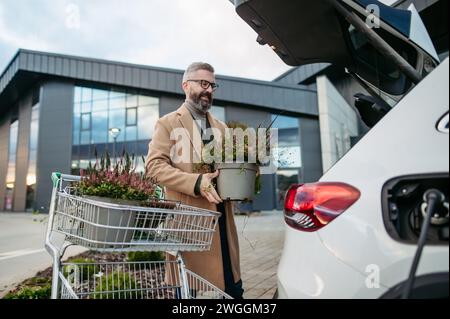 Man charging electric car before going to shopping. Public charging station in front of shop building, supermarket. Charging while shopping. Stock Photo