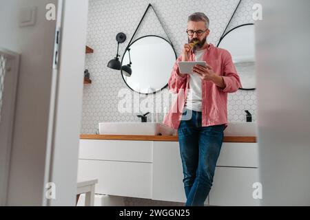 Man shopping online while brushing his teeth in bathroom. Holding tablet, scrolling and buying in eshop, toothbrush in mouth. Stock Photo