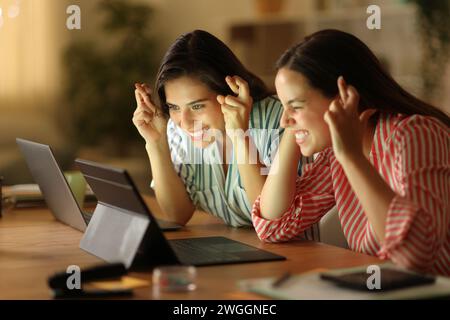 Two tele workers crossing fingers waiting for online result checking laptop in the night Stock Photo