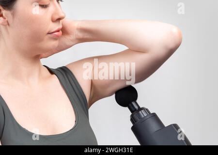 Close up of Caucasian woman gives herself massage on upper arm with percussion massager-gun with ball nozzle. Gray studio background. Therapy for join Stock Photo