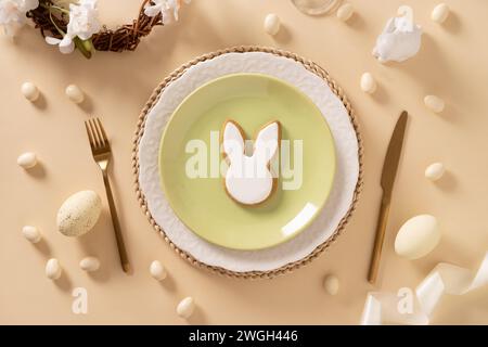 Easter table setting with white and green plate, bunny cookies, eggs on beige background. View from above. Happy Easter. Stock Photo