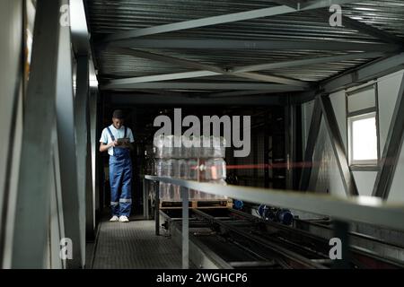 Young African American male engineer of bottled water factory using tablet while moving along conveyor belt in warehouse Stock Photo