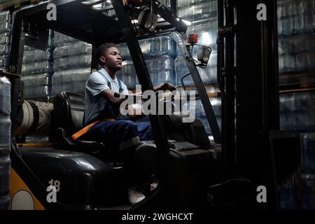Young African American man in workwear sitting in forklift and driving it along stacks of packages while holding by steer Stock Photo