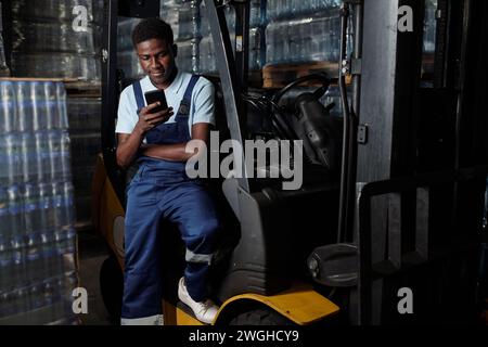 Young male engineer of industrial plant sitting by door of forklift and texting in mobile phone while having break after work Stock Photo