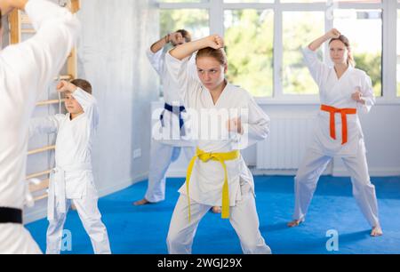 Girl with her family in kimonos and colored belts practicing karate punch block during group martial arts lesson in gym, accompanied by trainer Stock Photo
