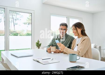 Middle aged happy couple using smartphone relaxing at living room table at home. Stock Photo