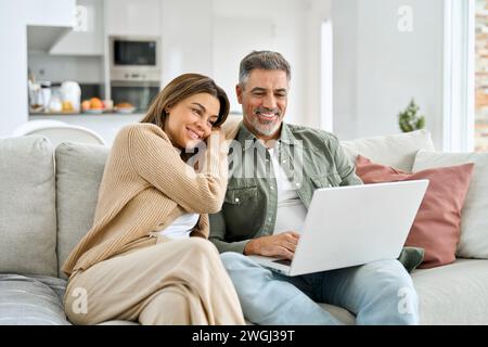 Happy middle aged couple using laptop relaxing on couch in living room. Stock Photo