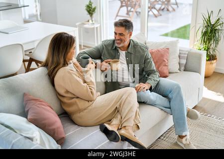Happy middle aged couple talking drinking coffee relaxing on couch at home. Stock Photo