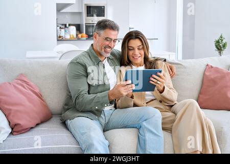 Happy middle aged couple using digital tablet relaxing on couch at home. Stock Photo