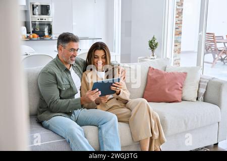 Happy middle aged couple using digital tablet relaxing on sofa at home. Stock Photo