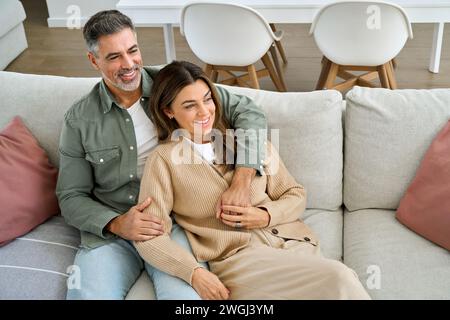 Happy middle aged mature couple hugging looking away relaxing on couch at home. Stock Photo