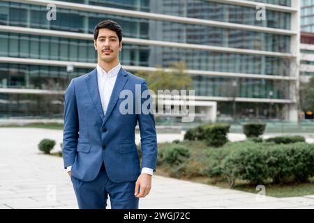 A confident Hispanic businessman stands in a relaxed pose outdoors with an urban backdrop, exuding professionalism and approachability in a tailored blue suit and crisp white shirt. Stock Photo